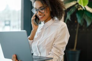 African businesswoman talking phone and holding laptop while standing near window in coworking